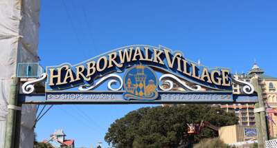 A blue sign reading "Harborwalk Village" spans over a street,...