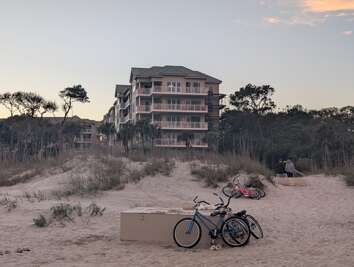 Bicycles rest on sandy dunes in front of a multi-story building...