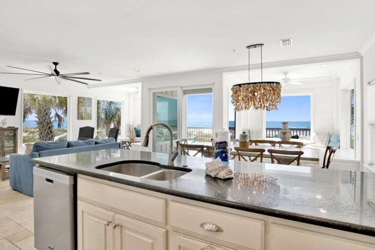 Black granite kitchen island with double sink overlooks dining area and ocean through sliding glass doors.