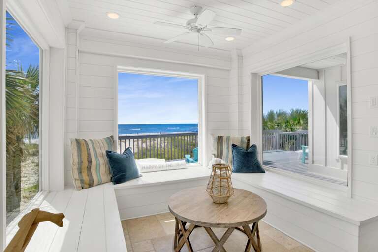 Bright white shiplap seating nook with striped and blue pillows, natural wood table, and ocean views through large windows.