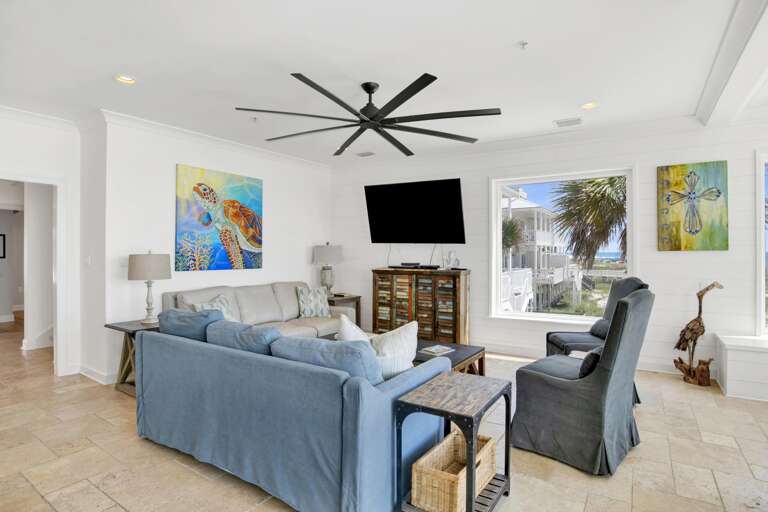 Living room with blue and beige sofas, high-back armchairs, rustic wood cabinet, and ocean views through large windows.