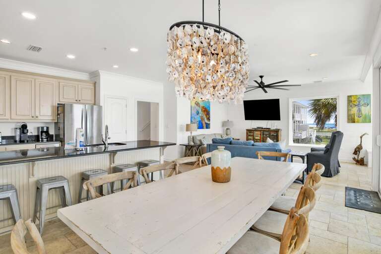 Large dining table with rustic wood chairs beneath a shell chandelier, adjacent to granite kitchen island and living area.