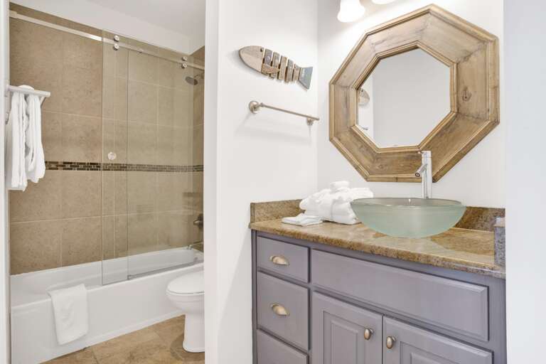 Bathroom with frosted glass vessel sink, granite countertop, octagonal wood-framed mirror, and tiled tub-shower combo.