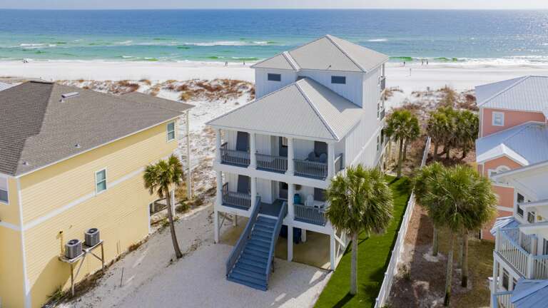 Exterior view of a white, three-story Gulf front house with blue railings, twin balconies, palm trees, and a sandy beach backdrop.