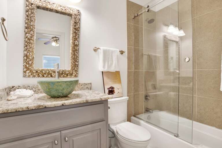 Bathroom with granite countertop, green vessel sink, seashell-framed mirror, and tub with glass sliding doors.