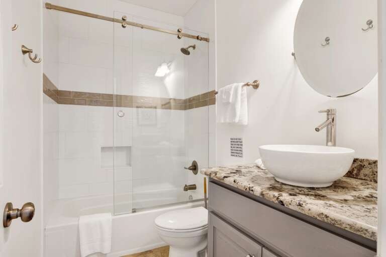 Bathroom with granite countertop, vessel sink, round mirror, and tub-shower combo behind sliding glass doors.
