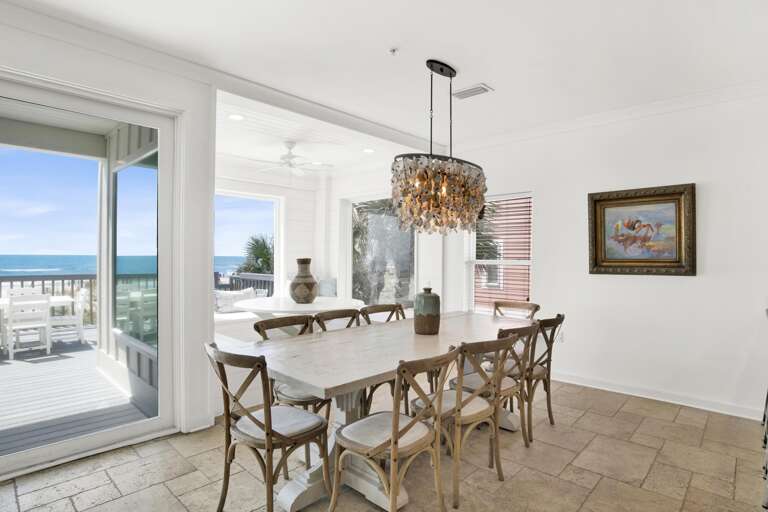 Dining room with weathered white wood table, eight cross-back chairs, shell chandelier, and ocean views through sliding doors.