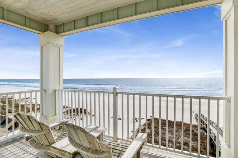 Wooden Adirondack chairs face the Gulf from a covered balcony with white railings and weathered wood flooring.