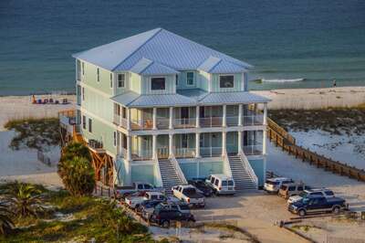 Three-story gulf front house with pale green siding, metal roof, double staircases, and sandy beach access ramp.