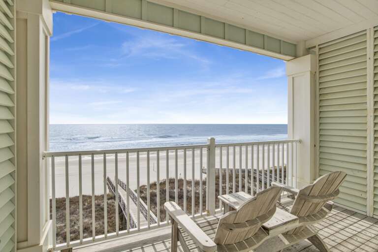Wooden Adirondack chairs face the ocean from a covered balcony with white railings and pale green siding.