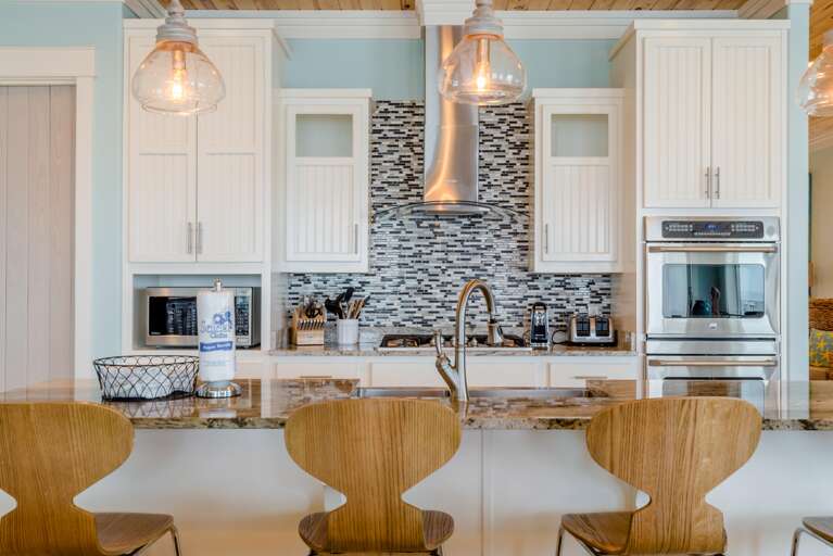 Kitchen island with granite countertop, three wood barstools, stainless steel sink, and mosaic tile backsplash.
