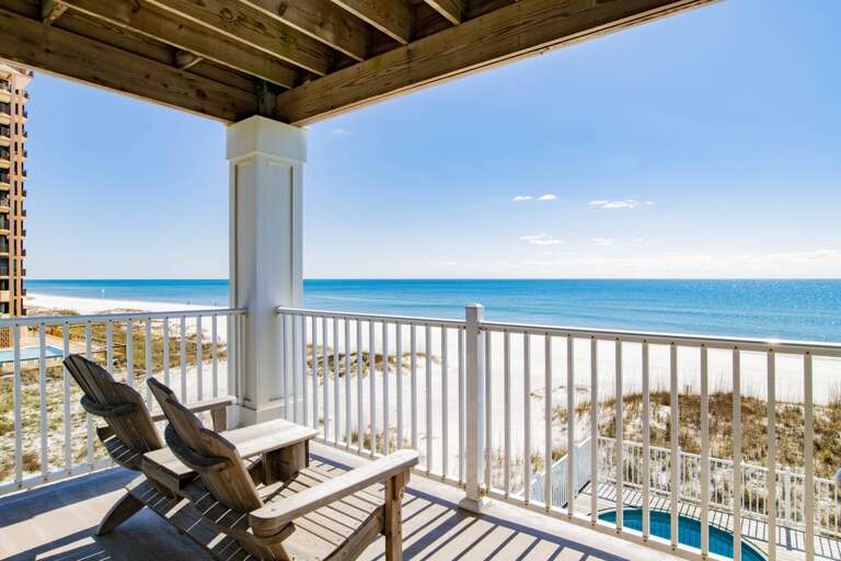 Covered balcony with white railing and two wooden Adirondack chairs overlooking white sand beach and blue Gulf waters.