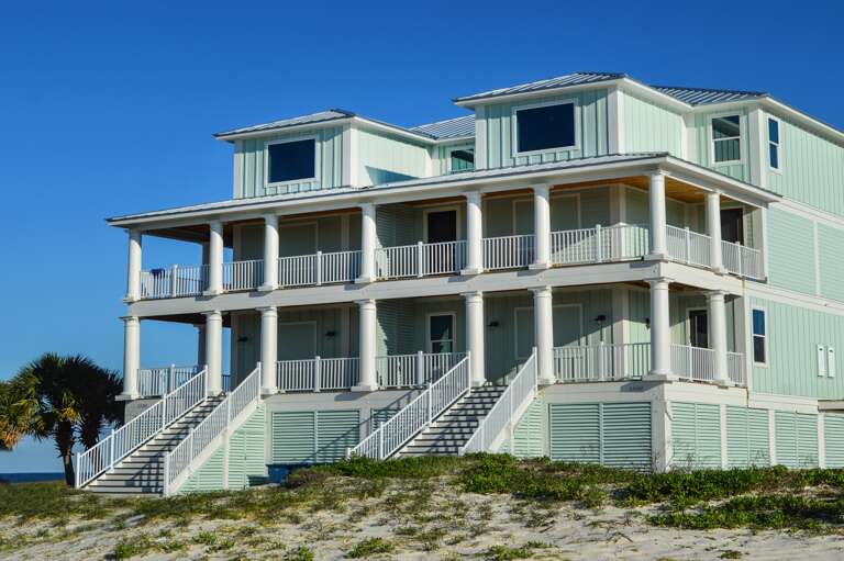 Two-story pale green gulf front house with white columns, metal roof, multiple staircases, and wraparound balconies.
