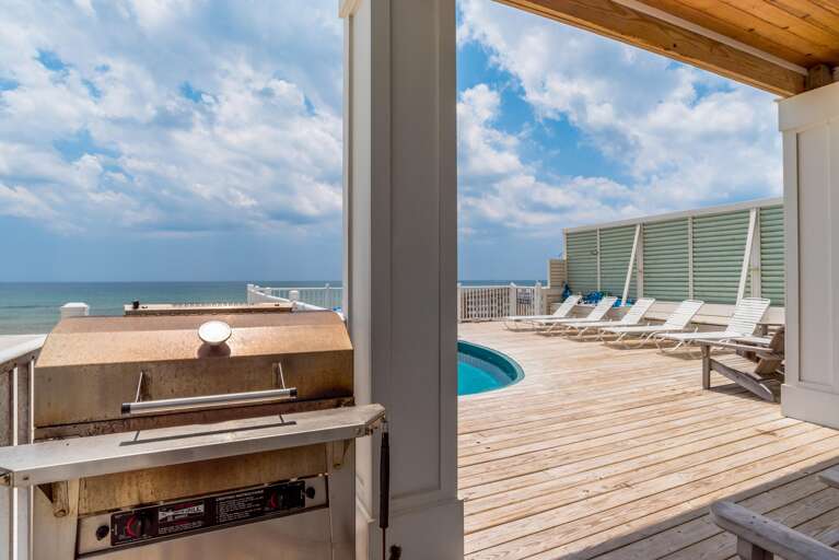 Weathered stainless steel gas grill positioned under a covered wooden roof, overlooking a wood-plank deck with white lounge chairs and pool.