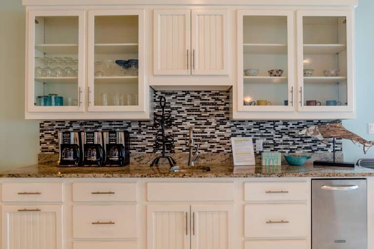 Kitchen counter with granite surface, mosaic tile backsplash, triple coffee makers, and glass-front white cabinets.
