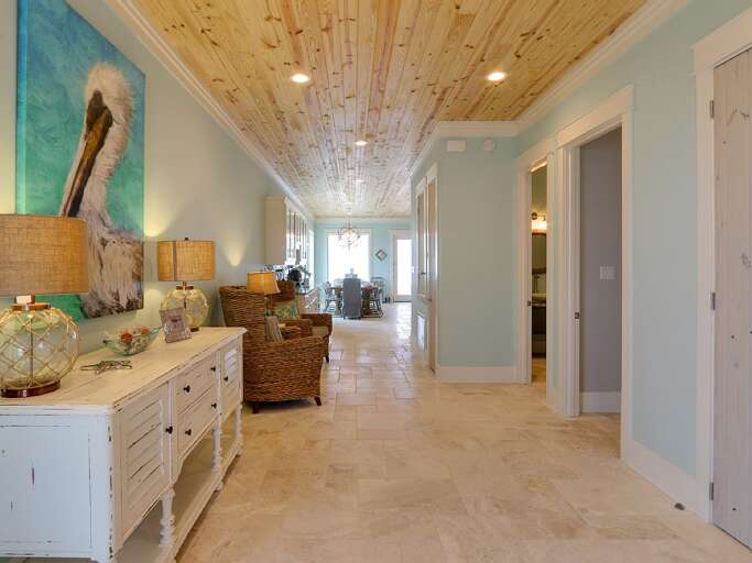 Long hallway with pale blue walls, natural wood plank ceiling, travertine tile floor, and wicker armchairs.
