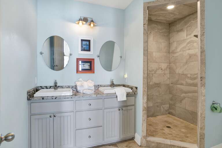 Bathroom with dual rectangular sinks, granite countertop, round mirrors, and a walk-in tile shower with recessed lighting.