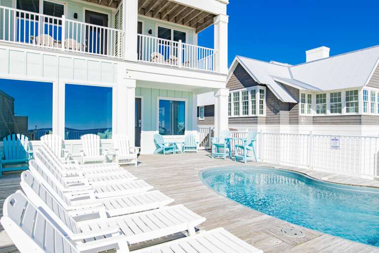 Curved pool edged by weathered wood decking with white lounge chairs and pastel blue Adirondack seating.
