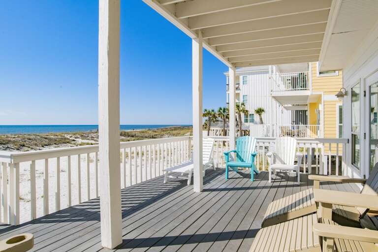 Spacious beachfront deck with gray wooden planks, Adirondack chairs in soft tan, white, and turquoise, shaded by a white pergola.