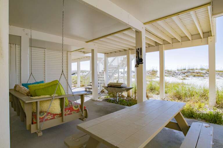 Shady ground-level patio with a cushioned wooden porch swing, picnic table, and grill facing sandy dunes.