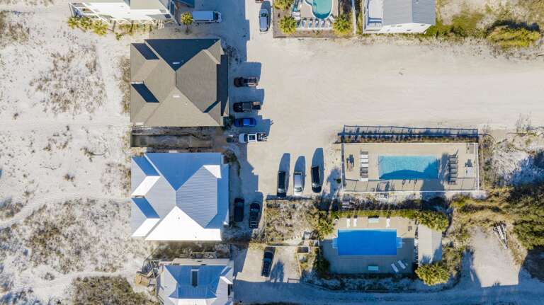 Overhead view of two fenced rectangular pools with lounge chairs, surrounded by sandy dunes and beach houses.