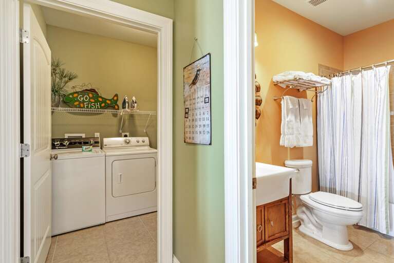 Laundry room with white washer and dryer, wire shelf with irons, adjacent to bathroom with striped shower curtain.