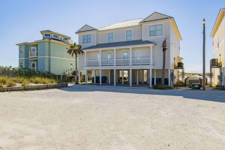 Raised white beach house with twin balconies, metal roof, and sandy parking area under clear blue sky.