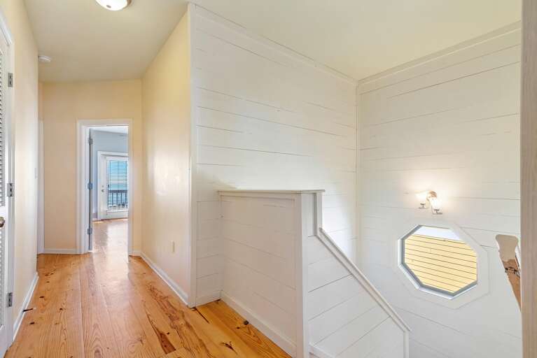 Hallway with honey-toned wood floors, shiplap walls, octagonal window, and a view into a bedroom with balcony doors.