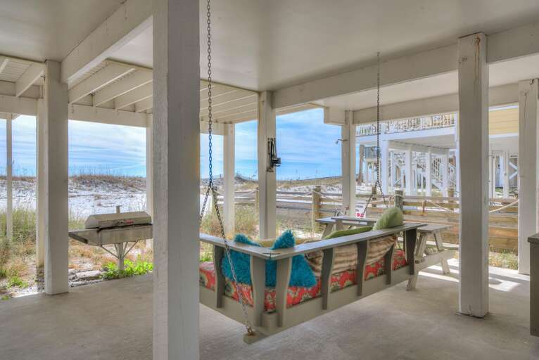 Covered patio with a colorful cushioned swing, picnic table, stainless steel grill, and sandy dunes beyond.