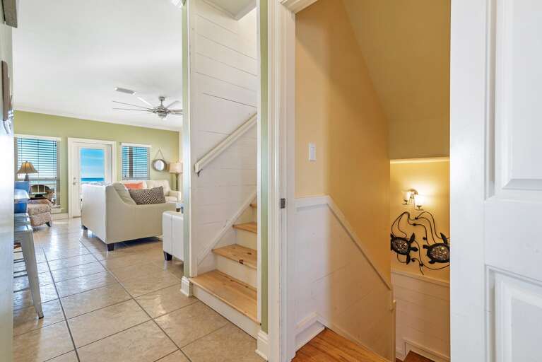 Entryway with natural wood stairs and tiled floor leads to a living room featuring cream sofas and ocean views through glass doors.