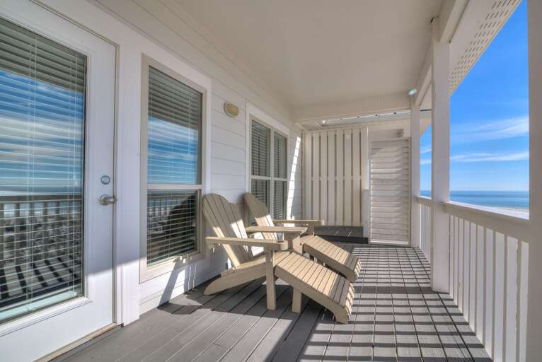 Sunlight casts shadows across the gray plank flooring and beige Adirondack chairs on the covered living room porch.