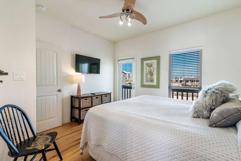Bedroom features light wood floors, a textured white bedspread, a wicker basket dresser, and a balcony with black railings.