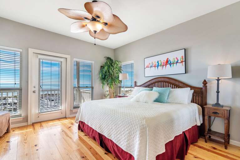 King bed with carved wood headboard, hardwood floors, and a ceiling fan shaped like palm leaves.
