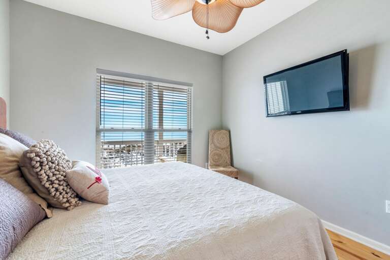 Bedroom with white quilted bedspread, textured pillows, woven chair, wall-mounted flat-screen TV, and window views.
