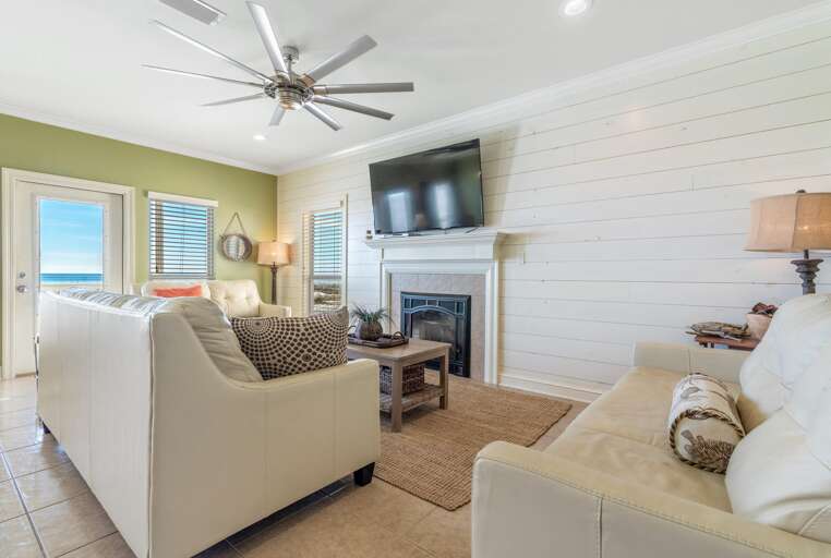 White leather seating arranged around a wooden coffee table on a woven rug, with tile flooring and a ceiling fan overhead.