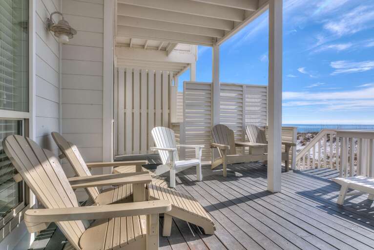 Deck features a mix of beige and white Adirondack chairs on weathered gray planks with ocean views beyond.