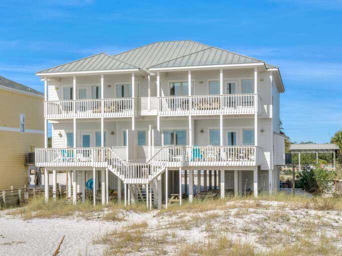Elevated white beach house with twin balconies, metal roof, and sandy dunes in the foreground under clear blue skies.