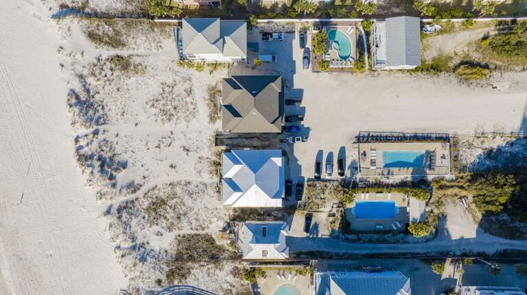 Aerial view of beachfront homes with metal roofs, sandy dunes, parked cars, and two fenced swimming pools.