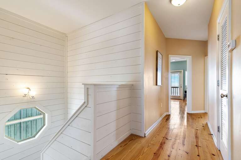 Hallway with honey-toned wood flooring, beige walls, white shiplap stairwell, and octagonal window near the staircase.