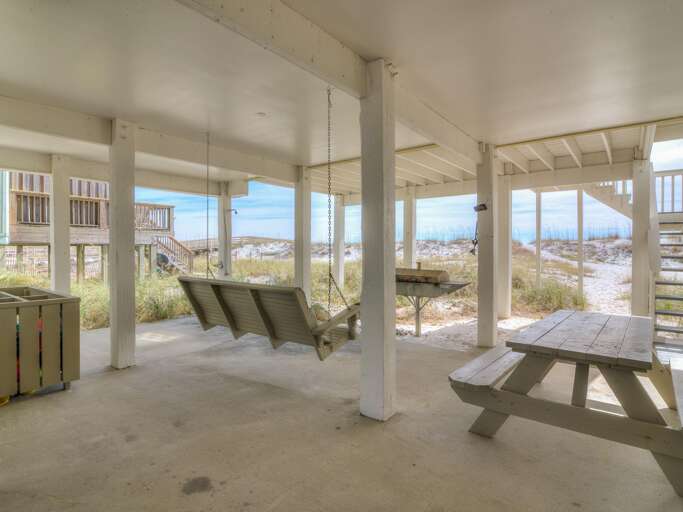 Concrete patio beneath the house with a hanging wooden porch swing, charcoal grill, and picnic table near dunes.