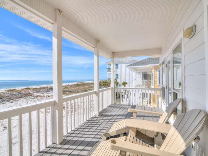 Living room opens to a covered porch with gray wood flooring, white railings, and tan Adirondack chairs facing the beach.