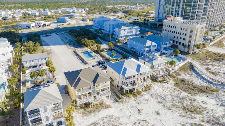 Sandy path leads from the elevated homes to beach dunes with sparse vegetation and footprints in the sand.