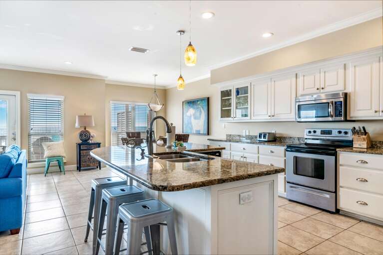 Granite-topped kitchen island with stainless steel sink and three metal stools beneath amber pendant lights.