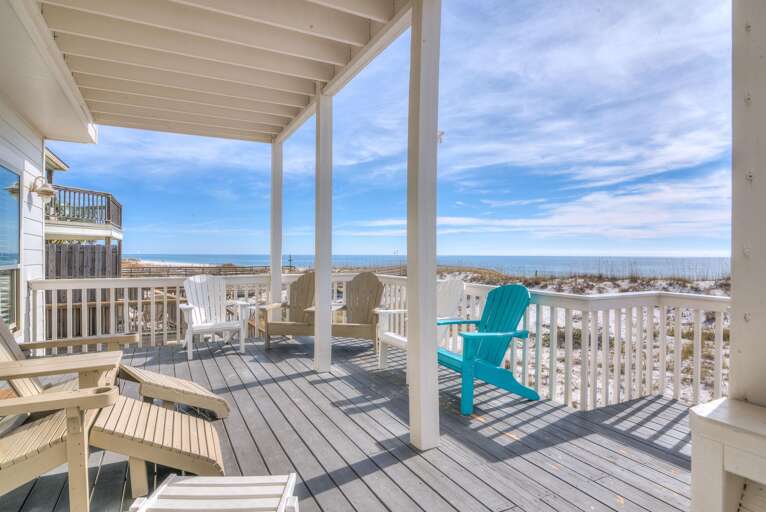 Covered porch with gray wood decking and white railings, Adirondack chairs, and views of sandy dunes and ocean.