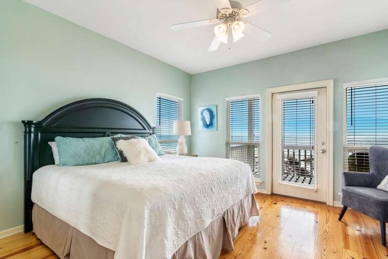 Bedroom with black arched headboard, light blue walls, hardwood floors, and a door leading to a balcony with ocean views.