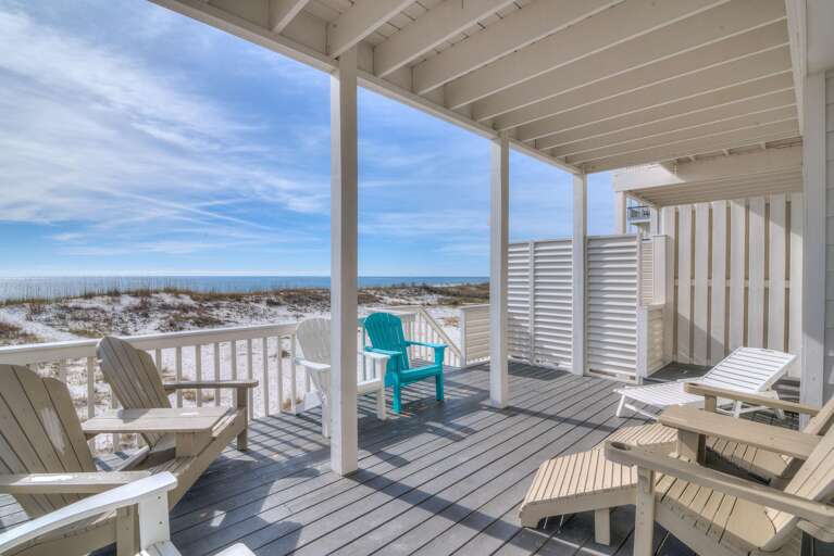 Covered beachside deck with gray wood flooring, Adirondack chairs, loungers, and ocean views past sandy dunes.