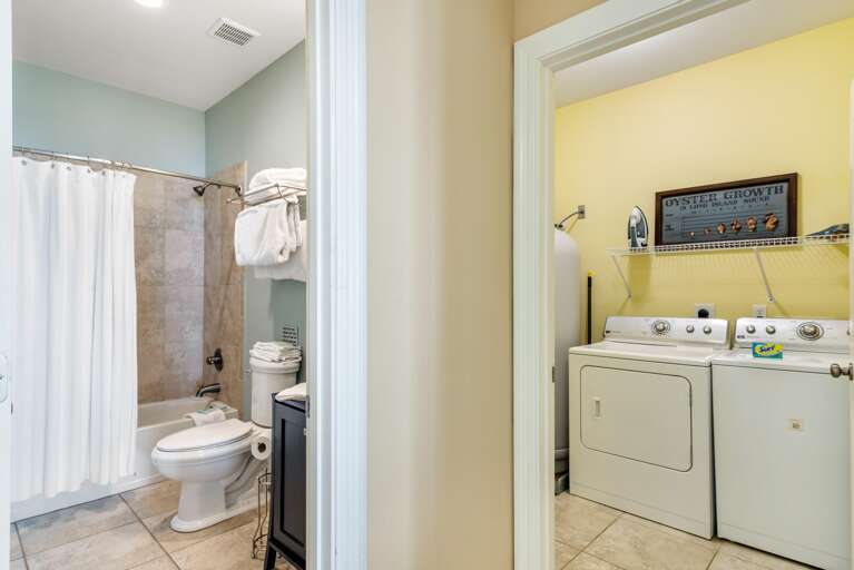 Bathroom with tiled tub surround, white shower curtain, and black vanity beside adjacent laundry room with washer and dryer.
