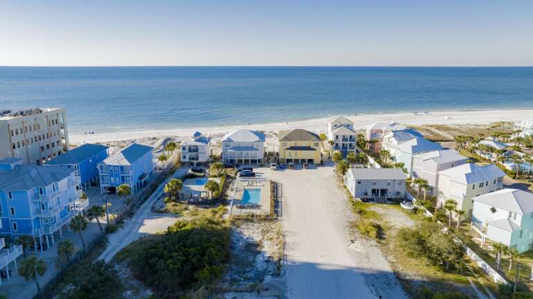 Wide exterior view shows a row of gulf-front homes with metal roofs, sandy driveways, and a fenced pool near the beach.