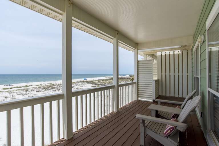 Covered balcony with dark wood flooring, gray Adirondack chairs, and white railings overlooking sandy dunes and ocean.
