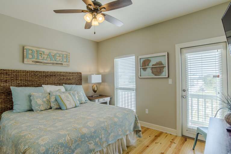 Bedroom features a woven headboard, coastal bedding, light wood floors, and a door leading to a balcony with blinds.